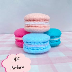 Op de afbeelding: Een stapel van vier vilten macaron-speeltjes in pastelroze, felroze, lichtblauw en turquoise, elk met een witte tussenlaag. Een roze wolk-vormige afbeelding in de linkerbenedenhoek bevat de zwarte tekst PDF Pattern.