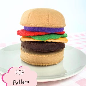 Könnte beinhalten: Ein Filz-Hamburger-Spielzeug mit beigen Brötchen, einem dunkelbraunen Patty, orangem Käse, grünem Salat, einer roten Tomatenscheibe und einem violetten Zwiebelring. Eine rosa Wolke zeigt den schwarzen Text PDF Pattern.