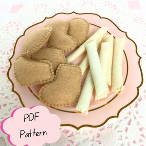 May include: A set of felt food items including tan heart-shaped pieces and off-white stick-shaped pieces arranged on a light pink plate with a gold rim. A pink cloud-shaped graphic with the text PDF Pattern is visible in the lower left corner.