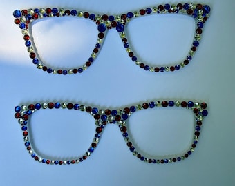 May include: Two pairs of eyeglass frames decorated with small, round, faceted rhinestones in alternating red, white, and blue colors. The frames are arranged horizontally, one above the other, featuring a classic cat-eye shape with clear outlines.
