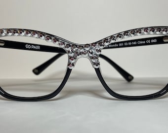 May include: Black cat-eye glasses with a clear lens and silver rhinestones around the frame. The glasses have the text "CO PAIR" on the left arm.
