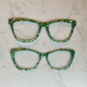 May include: Two pairs of green glitter cat eye glasses. The larger pair is on top of the smaller pair.