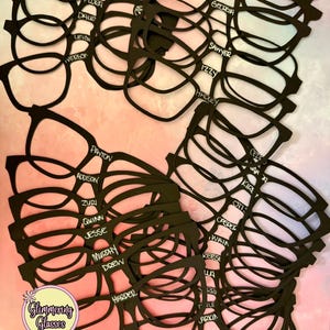 May include: A collection of black, flat, eyeglass-shaped cutouts, each featuring a unique name written in white script. The names include Evelyn, Flora, Dale, Payton, Addison, Zuri, Quinn, Jesse, Murphy, Drew, Harder, Breda, Nola, Sawyer, Izzy, Hadley, and others.