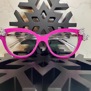 May include: Pink cat-eye glasses with clear lenses and silver rhinestone embellishments on the temples.