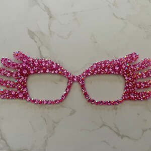 May include: Pink rhinestone glasses with a unique design. The glasses are made of small pink rhinestones that are arranged in a pattern that resembles a hand.