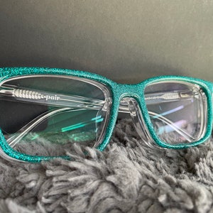 May include: Teal glitter square-shaped eyeglasses with clear lenses. The brand name "e-pair" is printed on the frame.