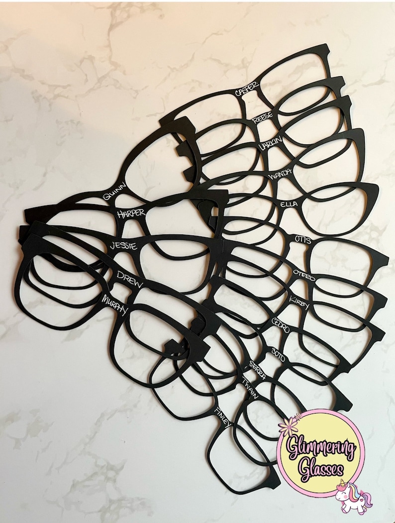 Puede incluir: Recortes de papel negro en forma de gafas con nombres escritos en ellos, incluyendo Reese, Quinn, Casper, Harper, Jessie, Drew, Murphy, Larkin, Wanda, Ella, Otis, Otero, Kirby, Cedro, Soto, Serra, Twain, Finley. Las gafas est&aacute;n dispuestas en forma de abanico con el texto "Glammering Glasses" y un gr&aacute;fico de unicornio en la parte inferior.