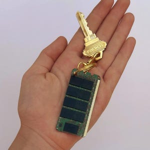 Puede incluir: Una llave dorada con la inscripción "SCHLAGE", unida a una memoria RAM de ordenador verde y negra mediante un llavero dorado. La llave y la memoria RAM se sostienen en una mano sobre un fondo blanco.