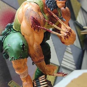 May include: A detailed figurine of a muscular character with dark hair, sideburns, and a cigar in their mouth. The figure wears a green tank top and shorts, features blood-stained skin, and has three sharp claws extended from each hand.