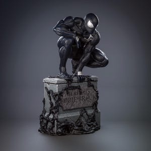 May include: A black symbiote figure in a crouching pose perched atop a grey stone tombstone. The tombstone features the engraved text HERE LIES EDDIE BROCK, with black, viscous, vine-like tendrils creeping over the stone surface and base.