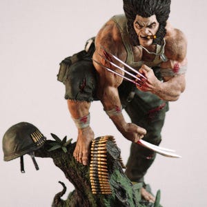 Peut inclure: Figurine détaillée de Wolverine, personnage de bande dessinée, griffes déployées. La figurine est posée sur un socle en forme de tronc d'arbre avec un casque militaire et une ceinture de munitions. La figurine porte un débardeur vert et un pantalon déchiré.