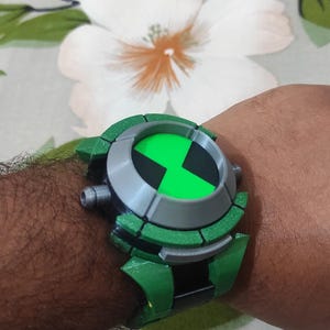 May include: A green and gray toy watch, resembling a popular cartoon series device, is shown on a person's wrist. The watch features a green face with a black and white design and a green band. The background is a floral pattern.