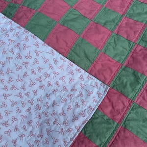 May include: A handmade quilt with a checkerboard pattern in shades of green and pink. The quilt's underside features a light blue fabric covered in a repeating pattern of pink bows. The quilt is likely made of cotton.