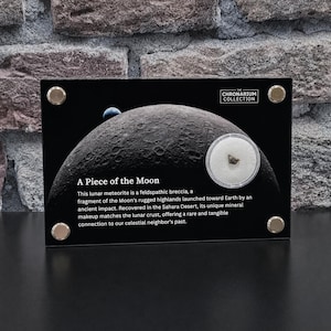 May include: A rectangular black display plaque titled The Chronarium Collection featuring a lunar meteorite fragment encased in a circular clear capsule. The plaque displays a graphic of the Moon and text describing the lunar feldspathic breccia.