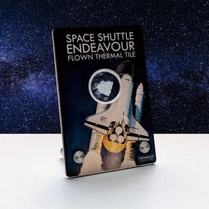 May include: A display piece featuring a Space Shuttle Endeavour thermal tile. The image shows the shuttle with flames and the text "SPACE SHUTTLE ENDEAVOUR FLOWN THERMAL TILE". The background is a starry night sky.
