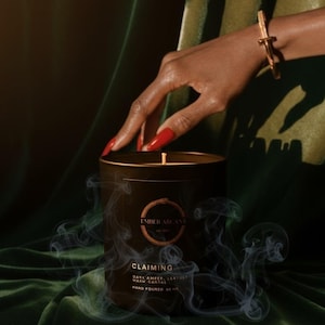 May include: A dark brown candle with the text "CLAIMING" and "EMBER ARCANA" is featured in this image. The candle is set against a dark green velvet backdrop, with wisps of smoke rising. A hand with red nail polish is reaching towards the candle.