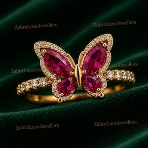 May include: A gold ring featuring a butterfly motif with four marquise-cut deep pink stones and small clear accent stones along the wings and band. The text ZahraLuxeJewellers is repeated across the image. The ring has a polished gold finish.