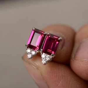 Può includere: Un paio di orecchini a bottone con pietre preziose rettangolari rosa intenso. Ogni orecchino ha un gruppo di piccole pietre chiare alla base, incastonate in un metallo color argento. Le pietre preziose hanno un aspetto vibrante e lucido.