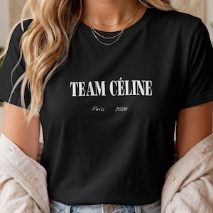 Peut inclure: Un t-shirt noir à manches courtes avec le texte blanc "TEAM CÉLINE" imprimé dans une police serif grasse, accompagné du texte plus petit "Paris 2026" centré juste en dessous. Le t-shirt présente un col rond classique et une coupe standard.