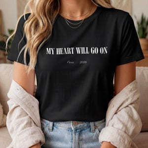 May include: A black short-sleeved t-shirt featuring the white text MY HEART WILL GO ON printed in a bold serif font, with the smaller white text Paris 2026 printed directly underneath the main phrase in a simple, clean sans-serif typeface.