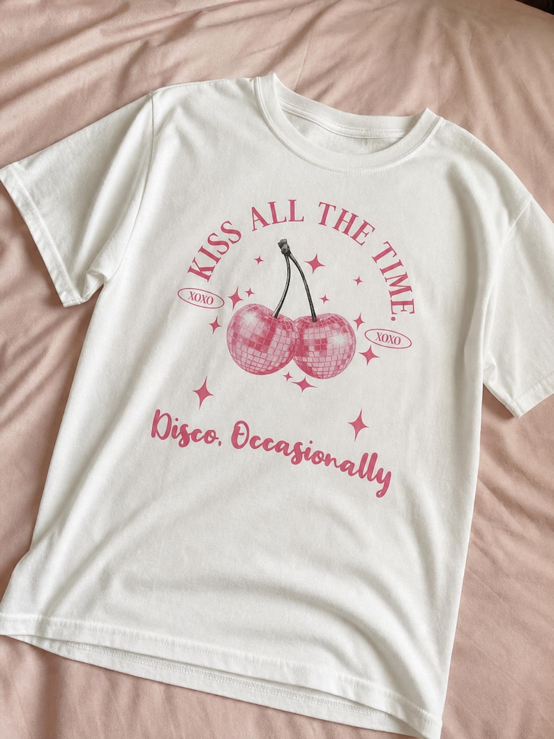 May include: White t-shirt with a pink graphic featuring two disco ball cherries and the text "KISS ALL THE TIME" and "Disco, Occasionally". The shirt has short sleeves and a crew neck.