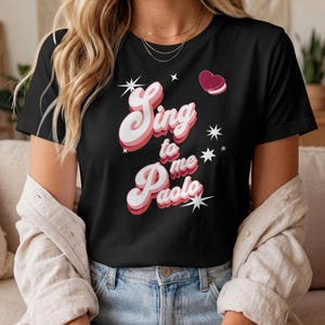 May include: A black short-sleeved t-shirt featuring the text "Sing to me Paolo" in a layered white and pink retro-style font. The design includes small white four-pointed star graphics and a single pink heart icon positioned above the text.