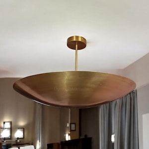 May include: A modern, brass-colored ceiling light fixture. The light has a circular, shallow bowl shape with a central stem and a round ceiling mount. The fixture is suspended from the ceiling and provides ambient lighting.