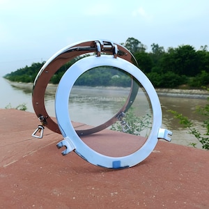 Puede incluir: Un ojo de buey cromado brillante con un panel de vidrio transparente, abierto y exhibido contra un fondo natural. El ojo de buey es circular con un mecanismo de apertura con bisagras. El texto "LightingByMantasha" es visible.