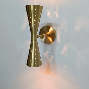 May include: A brass-toned wall sconce with a double cone design. Each cone has a series of small, evenly spaced holes that emit light. The sconce is mounted on a circular base, casting a warm glow on the wall.