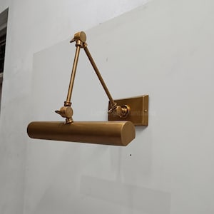 May include: A brass-toned wall-mounted picture light. The light has an adjustable arm and a cylindrical shade. The rectangular base is mounted on a white wall. The light fixture is designed to illuminate artwork.