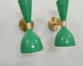 May include: Pair of mid-century modern wall sconces. Each sconce has a green, hourglass-shaped shade with white dot accents, a brass-coloured mounting plate, and a gold-coloured band. The sconces are mounted on a white wall.