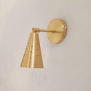 Puede incluir: Aplique de pared de latón con una pantalla cónica con pequeñas perforaciones. Tiene una placa trasera circular y un brazo recto. El aplique está montado en una pared clara.