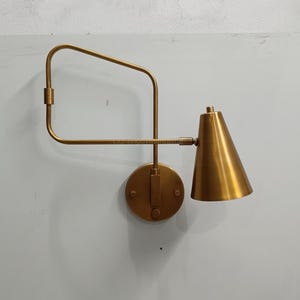 May include: A brass-colored wall-mounted lamp with a conical shade. The lamp features a circular base, a pivoting arm, and a rectangular frame. The design is minimalist and modern, suitable for interior lighting.