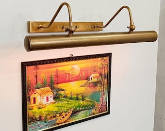 May include: A brass picture light fixture with a curved arm design, mounted above a framed landscape painting. The painting depicts a sunset scene with a river, houses, and trees. The text "Lighting By Master" is visible.