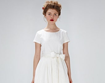 Puede incluir: Un vestido blanco con una cinta blanca y una pequeña flor roja en el pelo de la mujer.