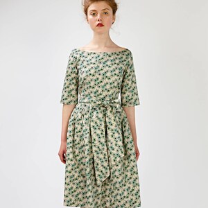 May include: A woman wearing a beige and green floral print dress with a matching sash tied at the waist. She is wearing brown heeled sandals with a bow detail.