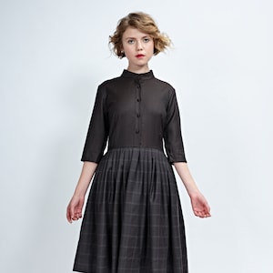 Può includere: Abito grigio scuro con collo alto e maniche a tre quarti. Il corpetto è grigio unito, mentre la gonna presenta un motivo a quadri. L'abito è abbinato a tacchi bordeaux, creando un look classico ed elegante.