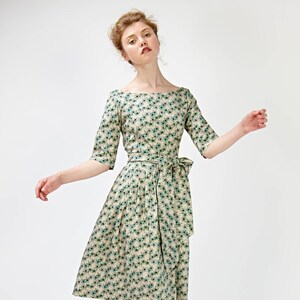 May include: A woman wearing a beige dress with a green floral pattern and a matching sash. She is wearing brown heeled sandals with a bow detail.