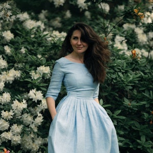 A woman in a light blue linen midi dress stands gracefully in front of a vibrant display of flowers.