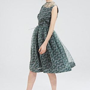 May include: A sleeveless, knee-length dress in a gray-green floral pattern. The dress features a sheer overlay and a matching belt. The model is wearing black high-heeled sandals with silver accents. The dress is suitable for a formal event.