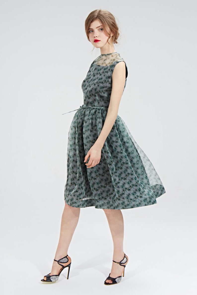 May include: A sleeveless, knee-length dress in a gray-green floral pattern. The dress features a sheer overlay and a matching belt. The model is wearing black high-heeled sandals with silver accents. The dress is suitable for a formal event.