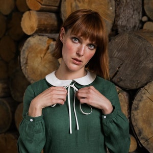 May include: A woman wearing a green dress with a white collar and a white bow. The dress has long sleeves and a gathered skirt. She is standing in front of a wall of stacked logs.
