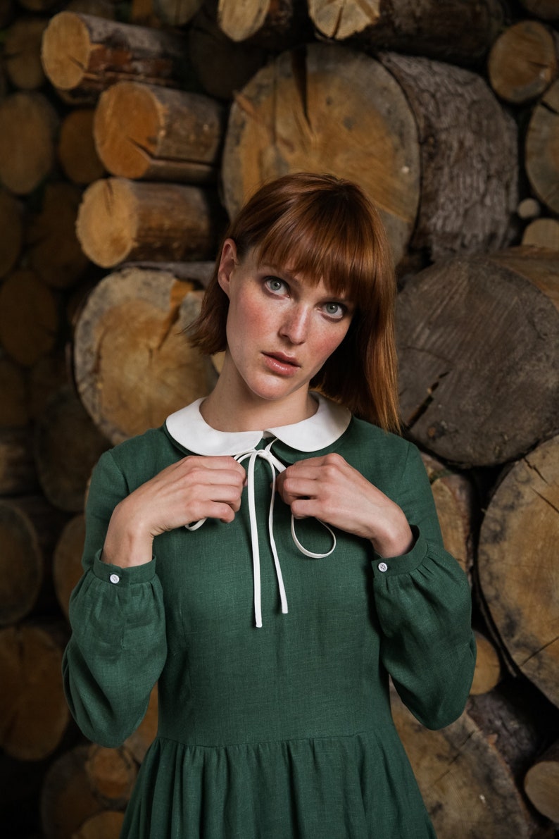 May include: A woman wearing a green dress with a white collar and a white bow. The dress has long sleeves and a gathered skirt. She is standing in front of a wall of stacked logs.
