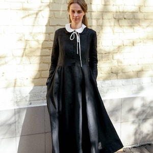 May include: A woman wearing a black linen dress with a white peter pan collar and white ribbon tie. The dress has long sleeves and a flowing skirt. She is standing in front of a brick wall.