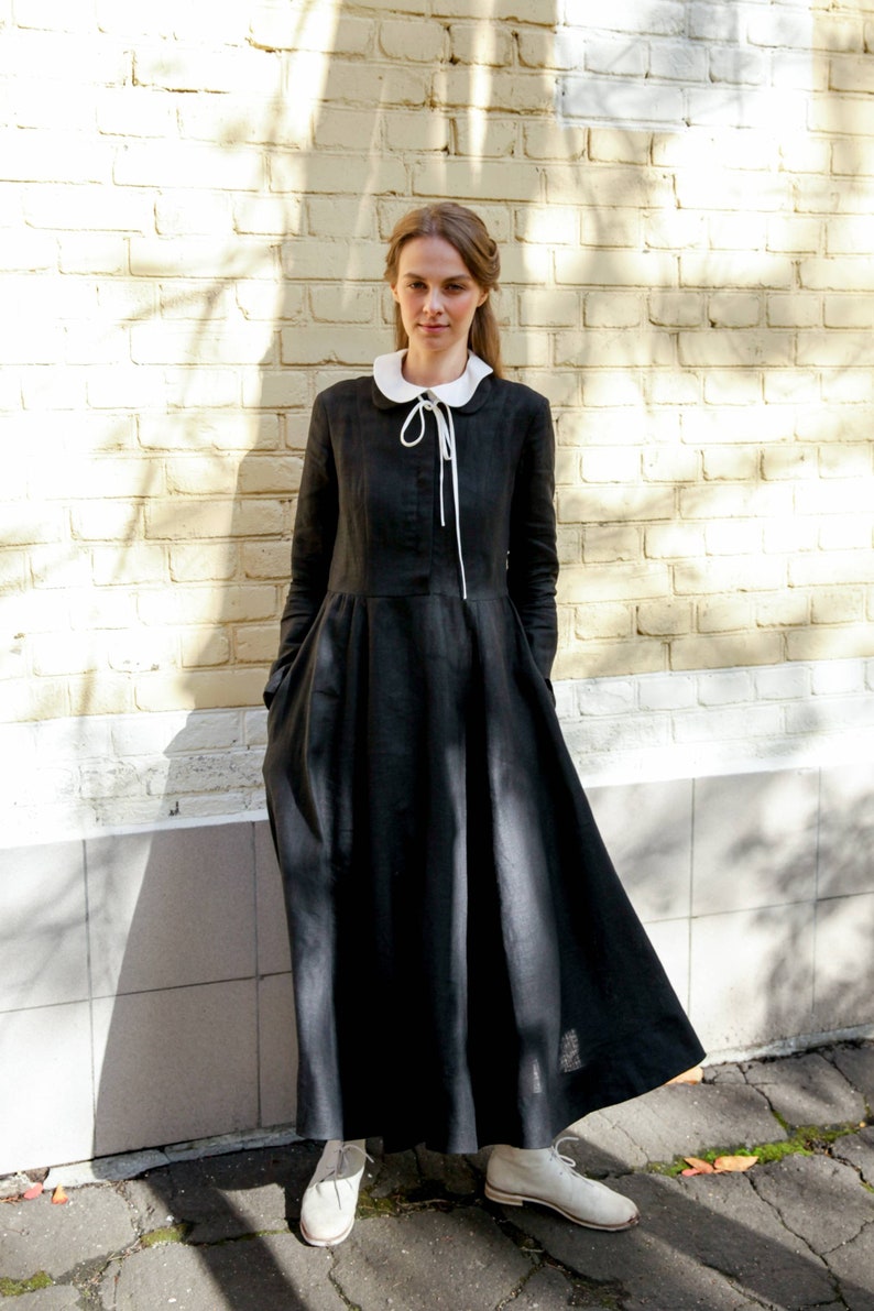 May include: A woman wearing a black linen dress with a white peter pan collar and white ribbon tie. The dress has long sleeves and a flowing skirt. She is standing in front of a brick wall.