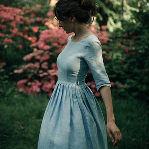 A woman in a light blue boatneck linen dress swirls in front of a lush garden
