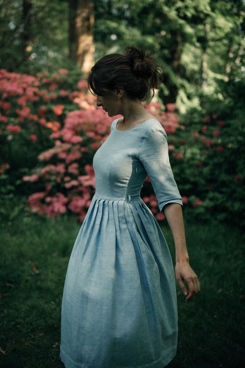 A woman in a light blue boatneck linen dress swirls in front of a lush garden