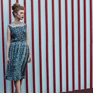 May include: A sleeveless, knee-length dress with a floral pattern and a sheer neckline. The dress is cinched at the waist with a thin belt. The background features a wall with alternating red and white vertical stripes.