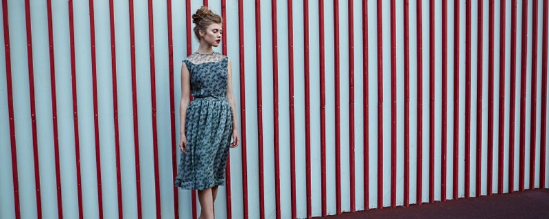 May include: A sleeveless, knee-length dress with a floral pattern and a sheer neckline. The dress is cinched at the waist with a thin belt. The background features a wall with alternating red and white vertical stripes.