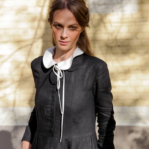 May include: A woman wearing a black linen dress with a white Peter Pan collar and a white ribbon tied at the neck. The dress has long sleeves and a slightly flared skirt. She is standing in front of a brick wall.